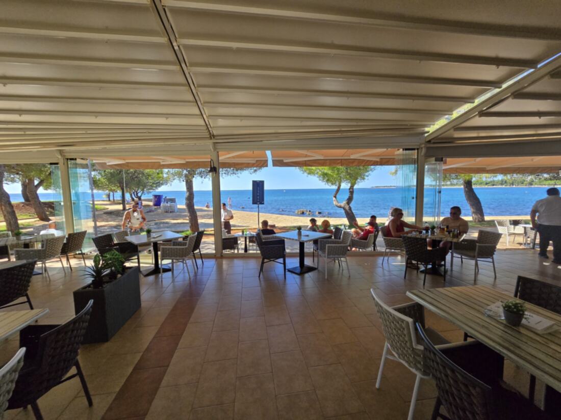Pod pergolom paviljona: stolovi iza staklene stijene i pogled na plažu i more