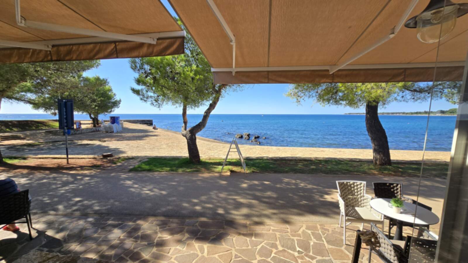 Pogled ispod karamel tende: dva nagnuta bora, šetnica, plaža i otvoreno more po sunčanom danu